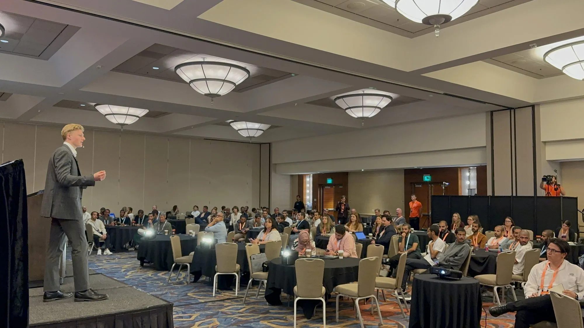 Speaker on stage presenting to a hotel ballroom full of conference attendees seated at round tables